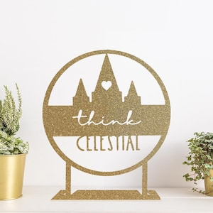 May include: Gold glitter decorative stand with a round top featuring a silhouette of a temple with a heart above it. The stand has a base and the words "think celestial" are written below the temple.