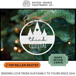 May include: White wooden ornament with a silhouette of a temple and the words "think celestial" hanging on a green Christmas tree.