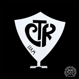 May include: White metal shield-shaped wall decor with a black CTR monogram and the name Liam below. The shield is mounted on a small white stand.