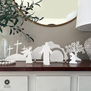 May include: White metal Easter scene featuring silhouettes of crosses, an angel, Jesus, a tomb, and a tree. The scene is displayed on a dark wooden surface. The logo for Rusted Orange is visible in the lower left corner.