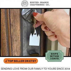 May include: White metal keychain shaped like a temple with a heart on top. The keychain is attached to a gold chain and is being held by a hand.