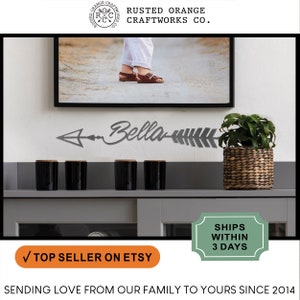 May include: A gray metal arrow with the name "Bella" written on it. The arrow is mounted on a wall above a shelf with four black candles. A potted plant is on the right side of the shelf. The text "RUSTED ORANGE CRAFTWORKS CO." is at the top of the image.