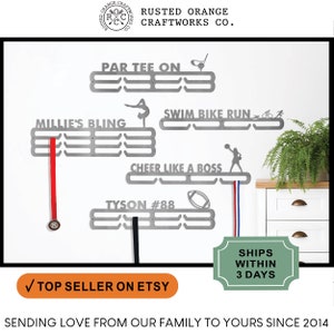 May include: A set of five silver medal hangers with various sports-themed designs. The hangers are mounted on a white wall with a green plant and a wooden cabinet in the background. The hangers feature the text "Par Tee On", "Swim Bike Run", "Millie's Bling", "Cheer Like A Boss", and "Tyson #88".