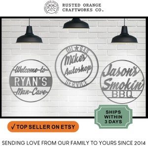 May include: Three round metal signs with custom text. The signs are silver and have black lettering. The signs read "Welcome to Ryan's Man-Cave", "Oil Gas Mike's Autoshop Service", and "Jason's Smokin' BBQ".