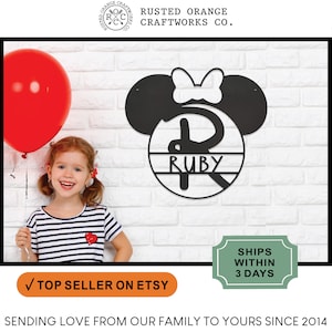 May include: Black metal Mickey Mouse ears with a white bow and a circle with the name "RUBY" inside. The ears are mounted on a white brick wall.