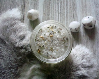May include: A glass jar filled with white bath salts, lavender, and other herbs. The jar is sitting on a wooden surface with white stones around it.