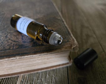 May include: A small amber glass roller bottle containing a golden liquid, featuring a white label with black text that reads "FOX & CRANE" and "THE FAMILIAR". The bottle has a clear plastic roller ball top and a separate black cylindrical cap.
