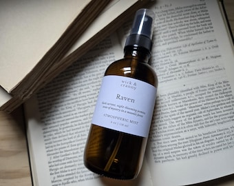 May include: An amber glass spray bottle with a black nozzle and a white label reading wick & cranny Raven black currant, night-blooming jasmine, sense of mystery in a moonlit forest ATMOSPHERIC MIST 4 oz | 118 ml, resting on an open book.
