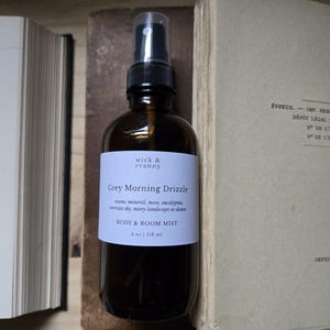 May include: A dark amber glass bottle with a black spray top, labeled "Grey Morning Drizzle" by Wick & Cranny. The label also states "BODY & ROOM MIST" and the volume is 4 fl oz / 118 ml. The bottle is positioned between two open books.