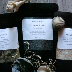 May include: Three small bags of bath salts with labels that read "Sea Witch Sea Soak", "Stormy Night Bath Soak", and "Moon Milk Bath". The bags are on a wooden surface with seashells and other natural elements.