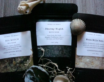 May include: Three small bags of bath salts with labels that read "Sea Witch Sea Soak", "Stormy Night Bath Soak", and "Moon Milk Bath". The bags are on a wooden surface with seashells and other natural elements.