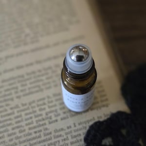 May include: A small, amber glass essential oil roller bottle with a silver rollerball and a white label. The bottle is resting on an open book with printed text. The background is blurred.