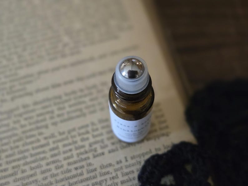 May include: A small, amber glass essential oil roller bottle with a silver rollerball and a white label. The bottle is resting on an open book with printed text. The background is blurred.