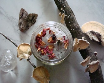 May include: A glass jar filled with white bath salts and dried rose petals. The jar is surrounded by various natural elements, including a branch with mushrooms, a moth, and a clear quartz crystal.