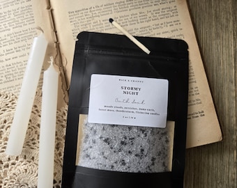 May include: A black pouch of Stormy Night bath salt with a white label reading Wick & Cranny, Stormy Night, Bath Salt, moody clouds, petrichor, damp earth, forest moss, thunderstorm, flickering candles, 2 oz | 56 g, featuring a single matchstick on top.