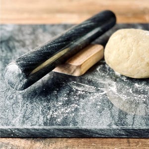 May include: A polished black marble rolling pin rests on a matching rectangular marble slab. A small wooden stand supports the rolling pin, and a ball of beige dough sits nearby on the stone surface, which is lightly dusted with white flour.