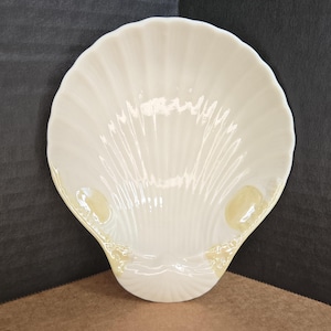 May include: A glossy, off-white ceramic dish shaped like a scallop shell. The surface features raised, radiating ridges that extend from the base to the scalloped edge, with subtle, light tan accents near the bottom hinge area.