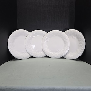 May include: A set of four round white ceramic plates arranged in a row. Each plate features a raised, embossed fruit and leaf pattern along the wide rim, while the center of each plate remains smooth, plain, and glossy white in appearance.