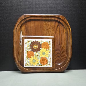 May include: A square wooden serving tray with rounded corners featuring a central square ceramic tile inlay decorated with a pattern of brown, orange, and yellow flowers. A small metal cheese knife with a wooden handle rests on the left side.