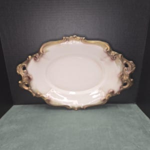 May include: An oval porcelain serving platter featuring a smooth white center and a decorative scalloped rim. The edges are detailed with ornate, raised scrollwork patterns finished in muted gold and soft brown tones, creating a textured border.