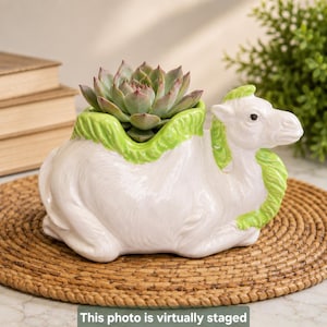 May include: A white ceramic camel-shaped planter with bright green accents on the saddle and neck area. The camel is in a seated position, and a green succulent plant is nestled into the opening on its back, serving as a decorative container.