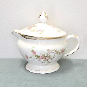 May include: A white porcelain sugar bowl with a lid, featuring a delicate floral pattern of pink roses and green vines. The bowl has a scalloped base, a curved handle, a small spout, and gold trim accents along the rim, lid, and handle edges.