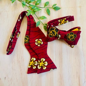 May include: A red bow tie with a yellow and white floral pattern. The bow tie has a matching adjustable strap.