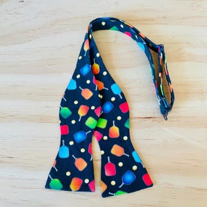 May include: A black bow tie with a colorful paddleball pattern. The bow tie has a black adjustable strap.