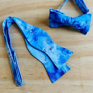 May include: Two blue bow ties with a marbled pattern. The bow ties are made of fabric and have shades of light and dark blue with white accents. One bow tie is tied, and the other is untied.