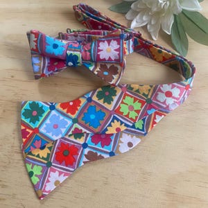 May include: A colorful bow tie and matching ascot set. The bow tie is pre-tied and features a vibrant floral pattern in shades of red, blue, green, and yellow. The ascot has a similar design, with a square pattern of flowers.