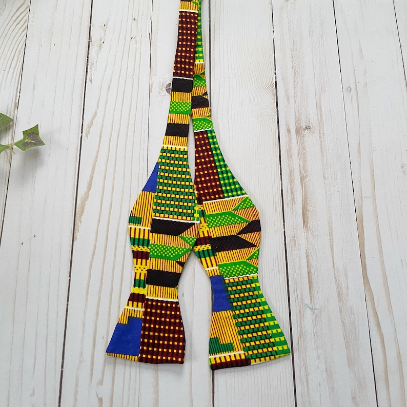 K&ouml;nnte beinhalten: Eine Fliege mit einem farbenfrohen afrikanischen Muster in Gelb-, Gr&uuml;n-, Braun- und Blaut&ouml;nen. Die Fliege ist aus Stoff gefertigt und hat ein traditionelles Design.