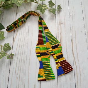 K&ouml;nnte beinhalten: Eine Fliege mit einem farbenfrohen afrikanischen Muster in Gelb-, Gr&uuml;n-, Blau- und Rott&ouml;nen. Die Fliege ist aus Stoff und hat ein traditionelles Design.
