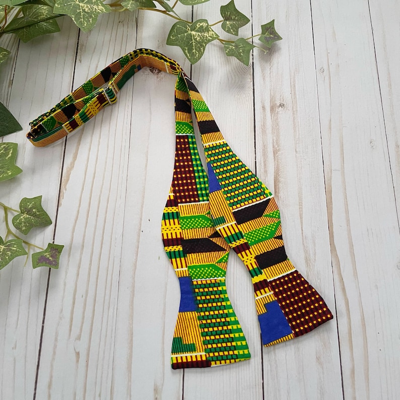 K&ouml;nnte beinhalten: Eine Fliege mit einem farbenfrohen afrikanischen Muster in Gelb-, Gr&uuml;n-, Blau- und Rott&ouml;nen. Die Fliege ist aus Stoff und hat ein traditionelles Design.