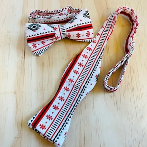 May include: A white bow tie and matching necktie set with a geometric pattern in red, black, and gray. The bow tie is pre-tied, and the necktie is adjustable. The set is displayed on a light wood surface.