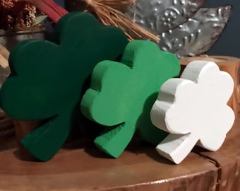 May include: Three wooden shamrock shapes, one green, one dark green, and one white. The shamrocks are arranged in a row on a wooden surface.