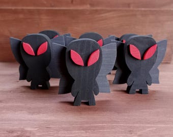May include: A group of wooden alien figurines. Each figurine is black with red eye shapes and gray wings. The figures are standing on a wooden surface.