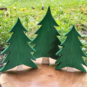 May include: Three wooden Christmas trees in various sizes, painted green, standing on a wooden surface. The trees are all in a similar style, with a simple, classic design.