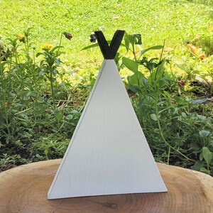 May include: White wooden teepee with a black V-shaped top. The teepee is sitting on a wooden surface.