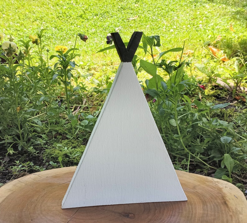 May include: White wooden teepee with a black V-shaped top. The teepee is sitting on a wooden surface.
