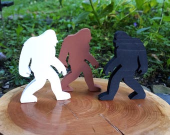 May include: Three standing figures shaped like Bigfoot, each in a different solid color. One figure is white, one is brown, and one is black. The figures are stylized with a side profile view, showing a hunched posture and long arms.