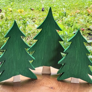 May include: Three wooden Christmas trees of varying sizes, painted green, stand on a wooden base. The trees are arranged in a semi-circle.