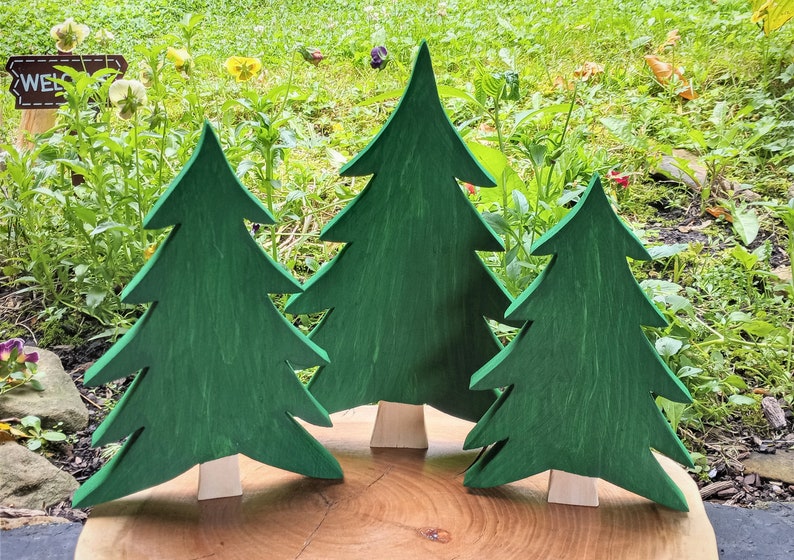 May include: Three wooden Christmas trees of varying sizes, painted green, stand on a wooden base. The trees are arranged in a semi-circle.