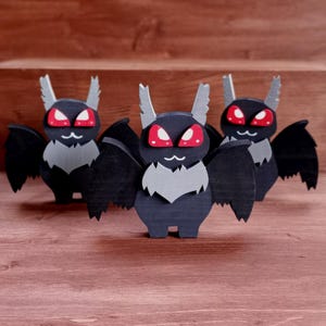 May include: Three wooden bat figurines with black bodies, gray wings and chest, and red eyes. The bats have white, pointed ears and are standing on a wooden surface. The background is a wooden box.