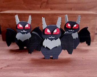 May include: Three wooden bat figurines with black bodies, gray wings and chest, and red eyes. The bats have white, pointed ears and are standing on a wooden surface. The background is a wooden box.