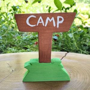 May include: A wooden sign with the word "CAMP" painted in white on a brown background. The sign is attached to a brown wooden post with a green base.