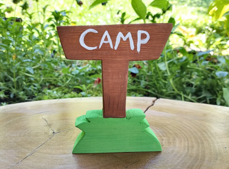 May include: A wooden sign with the word "CAMP" painted in white on a brown background. The sign is attached to a brown wooden post with a green base.