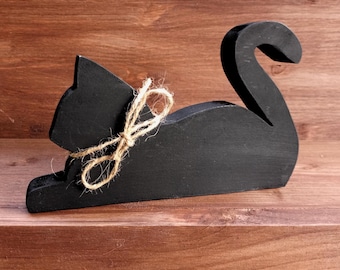 May include: Black wooden cat figurine with a brown twine bow. The cat is lying down with its tail curled around its body.