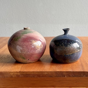 May include: Two small, round ceramic vases. One vase is a mix of pink, green, and tan colors. The other vase is dark blue with brown and purple hues. Both vases have a glossy finish and sit on a wooden table.