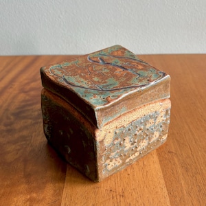 May include: A square ceramic box with a lid. The lid is a mix of brown, teal, and orange, with a textured surface. The box's sides are a speckled tan and gray. The box is sitting on a wooden surface.