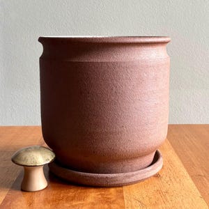 May include: A large, reddish-brown ceramic planter with a matching saucer. A small, mushroom-shaped decorative object sits to the left of the planter. The planter has a textured surface and a wide opening. The items are on a wooden surface.